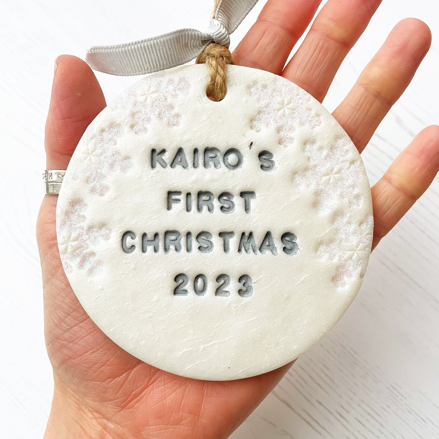 Personalised baby’s first Christmas bauble ornament, pearlised white clay round personalised with Kairo’s first Christmas 2023 in grey paint, decorated with 3 iridescent glitter snowflakes on either side of the bauble