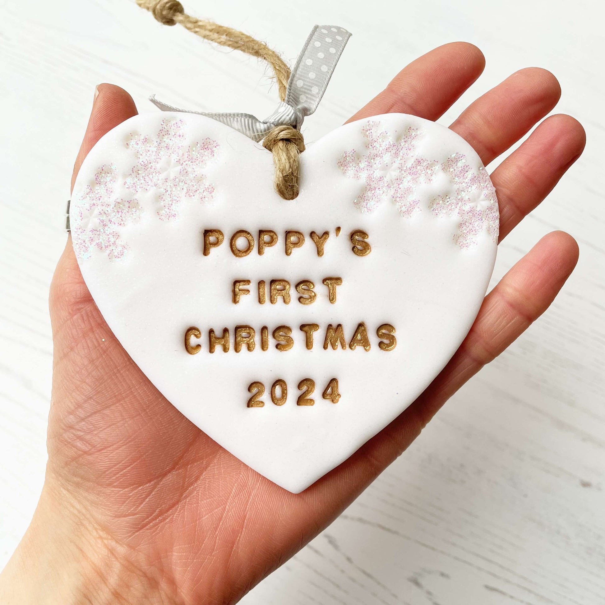 Personalised baby’s first Christmas heart ornament, glitter white clay with POPPY’S FIRST CHRISTMAS 2024 painted gold, decorated with 2 iridescent glitter snowflakes on either side of the top of the heart