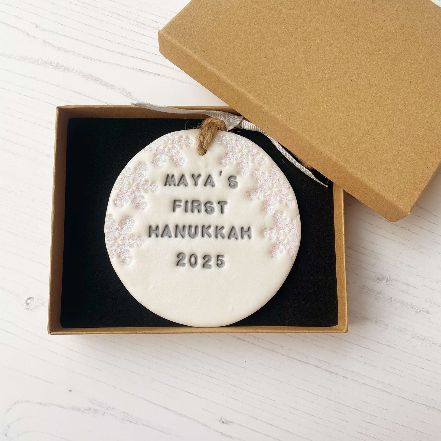 Personalised baby's first Hanukkah bauble ornament, pearlised white round clay with MAYA’S FIRST HANUKKAH 2025 in grey paint, decorated with 3 iridescent glitter snowflakes on either side of the bauble