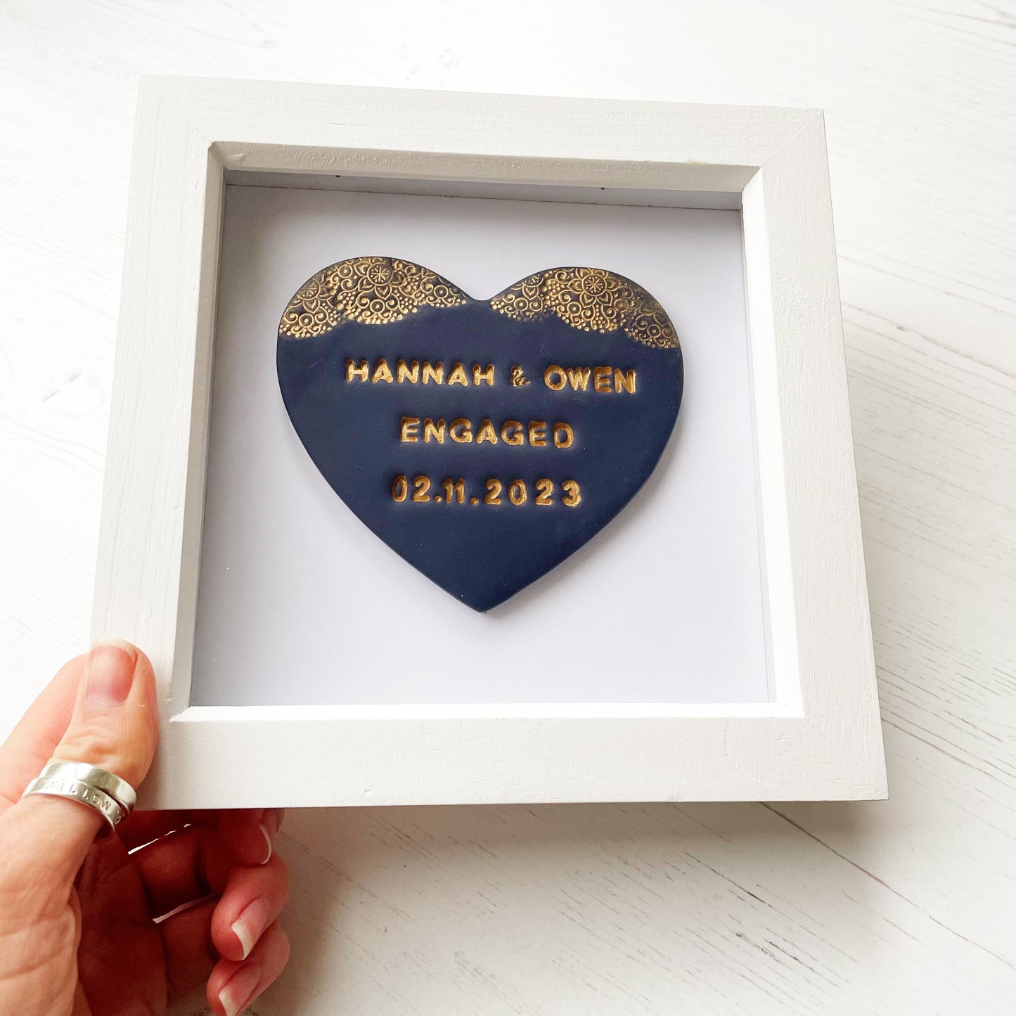 Personalised framed engagement gift, dark blue clay heart with a gold lace edge at the top of the heart in a white box frame, the heart is personalised with HANNAH & OWEN ENGAGED 02.11.2023