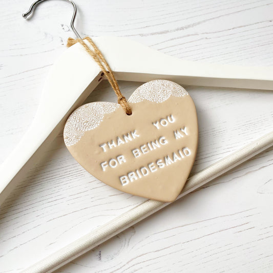 Personalised Bridesmaid thank you gift, beige clay hanging heart with a white lace edge at the top of the heart, hanging off a white coat hanger,  the heart is personalised with THANK YOU FOR BEING MY BRIDESMAID