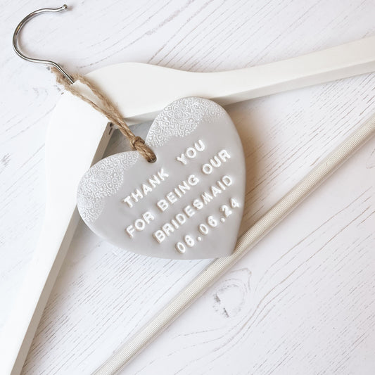 Personalised Bridesmaid thank you gift, grey clay hanging heart with a white lace edge at the top of the heart, hanging off a white coat hanger, the heart is personalised with THANK YOU FOR BEING MY BRIDESMAID 08.06.24