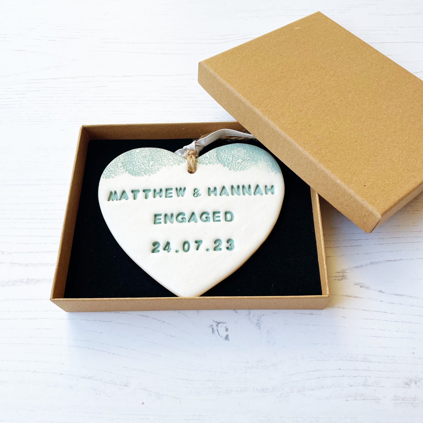 Personalised engagement gift, pearlised white clay hanging heart with a sage green lace edge at the top of the heart, the heart is personalised with MATTHEW & HANNAH ENGAGED 24.07.23 In a luxury Kraft brown gift box