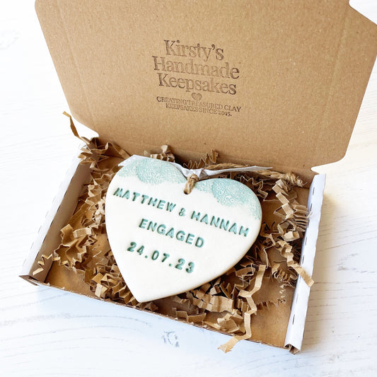 Personalised sage green engagement gift, pearlised white clay hanging heart with a sage green lace edge at the top of the heart, the heart is personalised with MATTHEW & HANNAH ENGAGED 24.07.23. In a white postal box with brown Kraft zigzag shredded paper.
