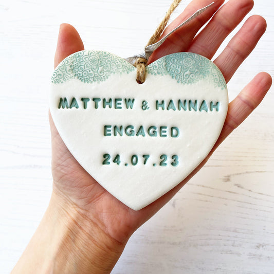 Personalised engagement gift, pearlised white clay heart with a sage green lace edge at the top of the heart with jute twine for hanging, the heart is personalised with MATTHEW & MANNAH ENGAGED 24.07.23