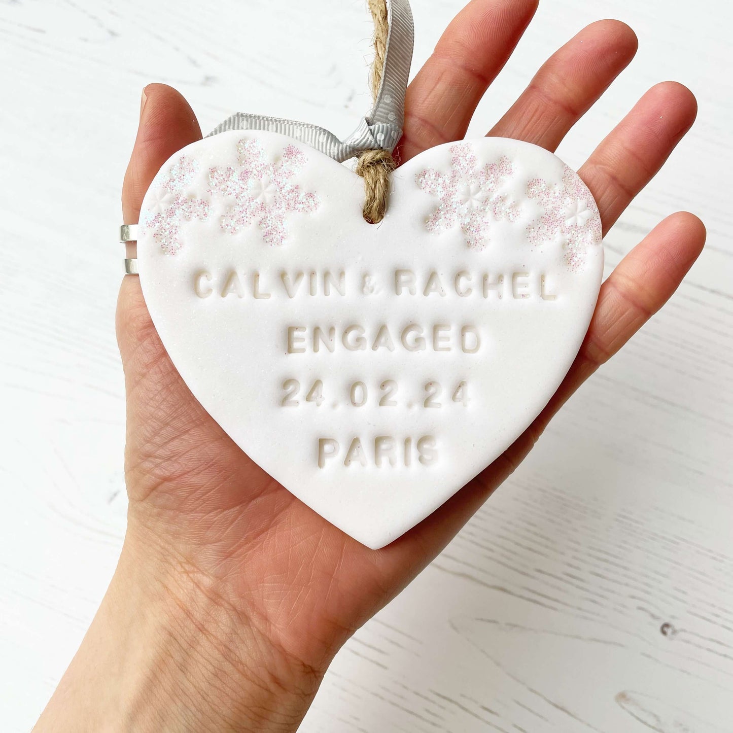Personalised first Christmas engaged heart ornament, glitter white clay with CALVIN & RACHEL ENGAGED 24.02.24 PARIS (text not painted), decorated with 2 iridescent glitter snowflakes on either side of the top of the heart
