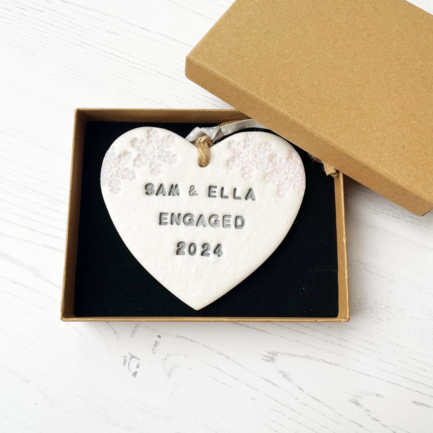 Personalised first Christmas engaged heart ornament, pearlised white clay with SAM & ELLA ENGAGED 2024 painted grey, decorated with 2 iridescent glitter snowflakes on either side of the top of the heart