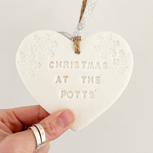 Personalised baby's first Christmas heart ornament, pearlised white clay with CHRISTMAS AT THE POTTS' (text not painted), decorated with 2 iridescent glitter snowflakes on either side of the top of the heart