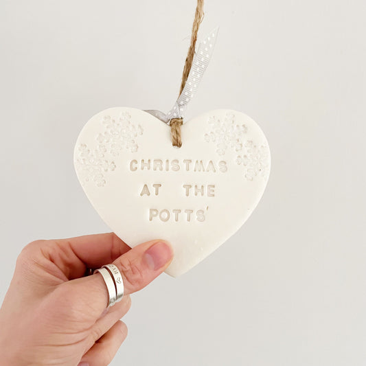 Personalised baby's first Christmas heart ornament, pearlised white clay with CHRISTMAS AT THE POTTS' (text not painted), decorated with 2 iridescent glitter snowflakes on either side of the top of the heart