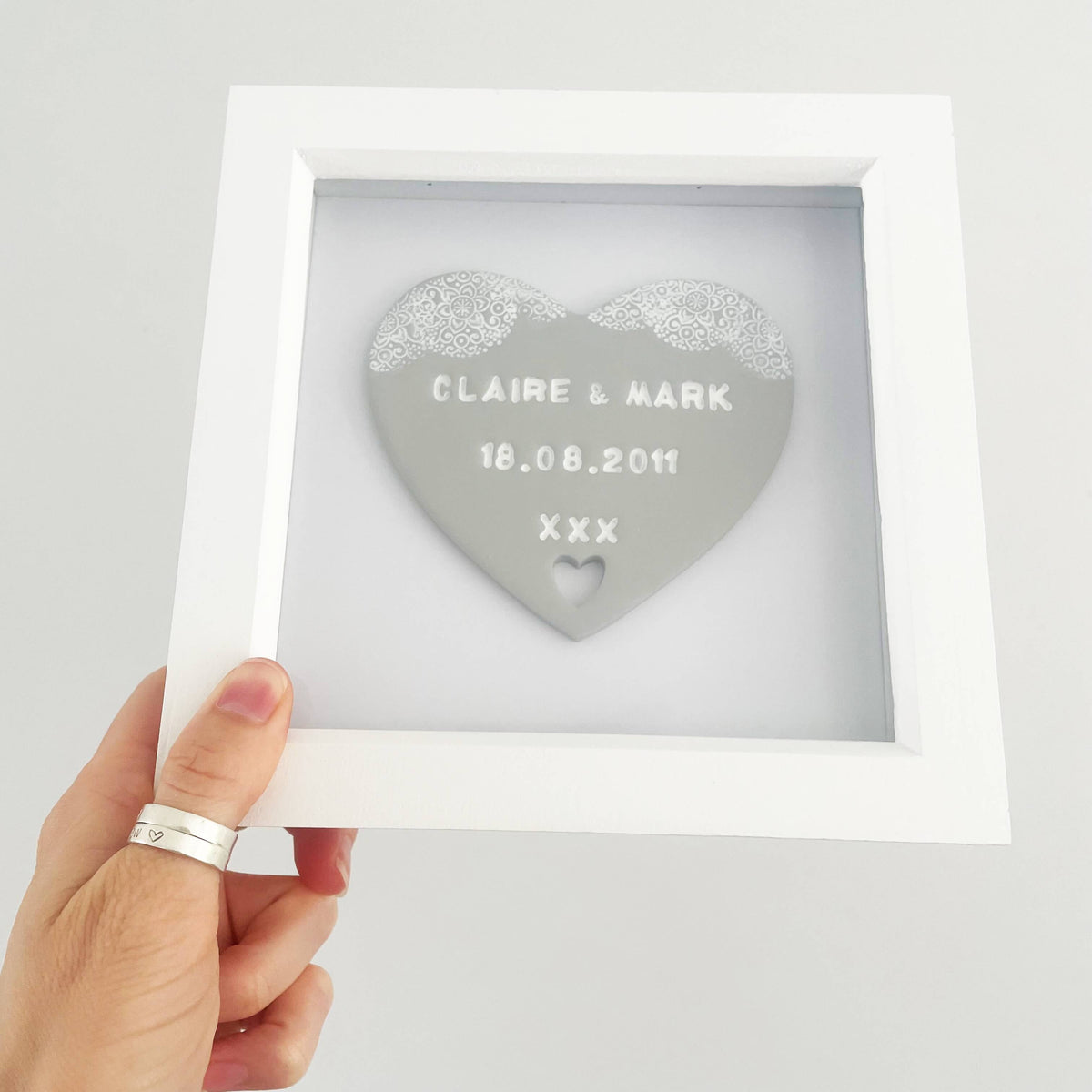 Personalised anniversary gift, grey clay heart with a white lace edge at the top of the heart and a heart cut out at the bottom in a white box frame, the heart is personalised with CLAIRE & MARK 18.08.2011 XXX