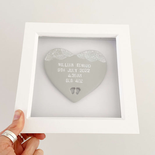 Personalised baby keepsake gift, grey clay heart with baby feet cut out at the bottom of the heart and a white lace edge at the top of the heart in a white box frame, the heart is personalised with the baby’s name, date of birth, weight and time