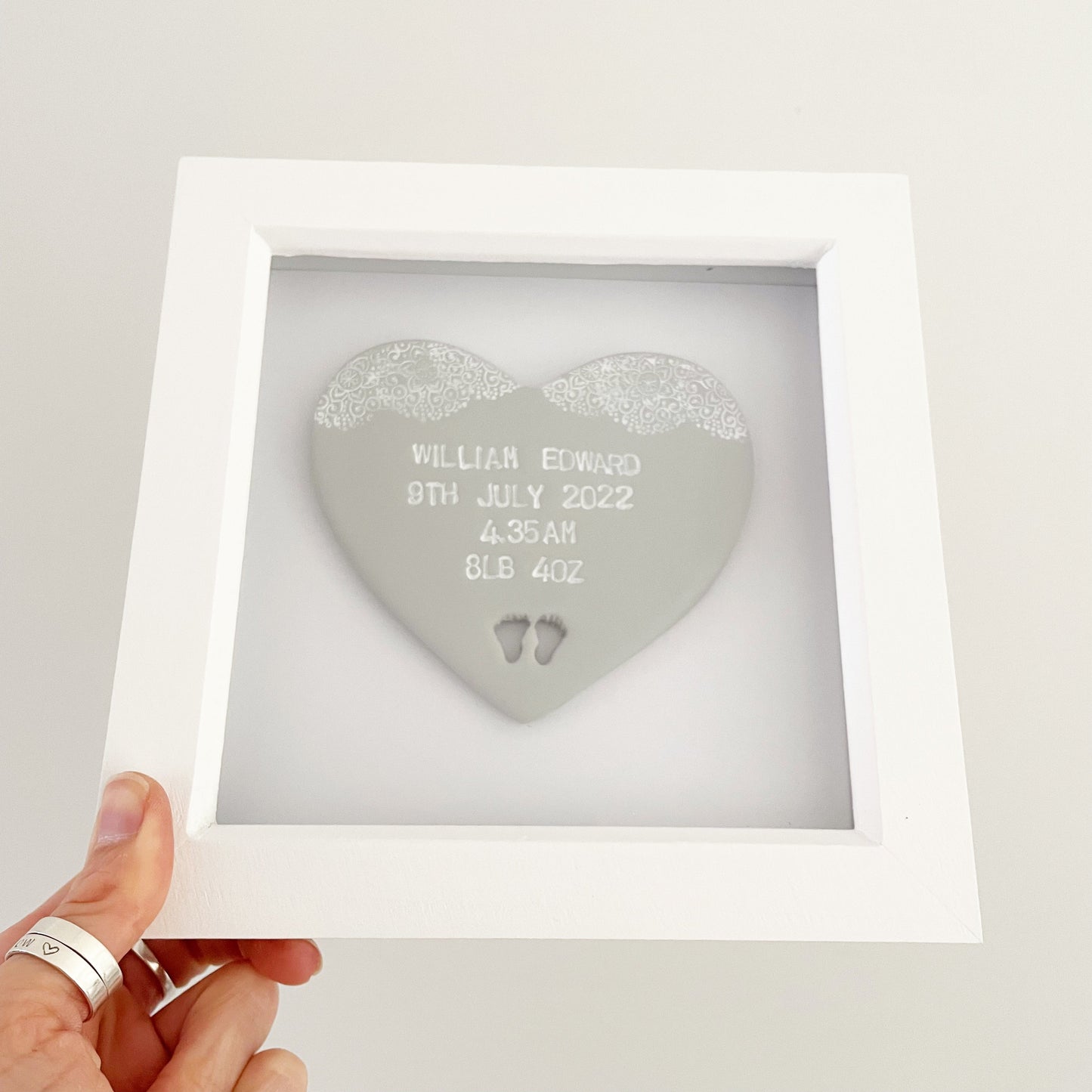 Personalised baby keepsake gift, grey clay heart with baby feet cut out at the bottom of the heart and a white lace edge at the top of the heart in a white box frame, the heart is personalised with the baby’s name, date of birth, weight and time