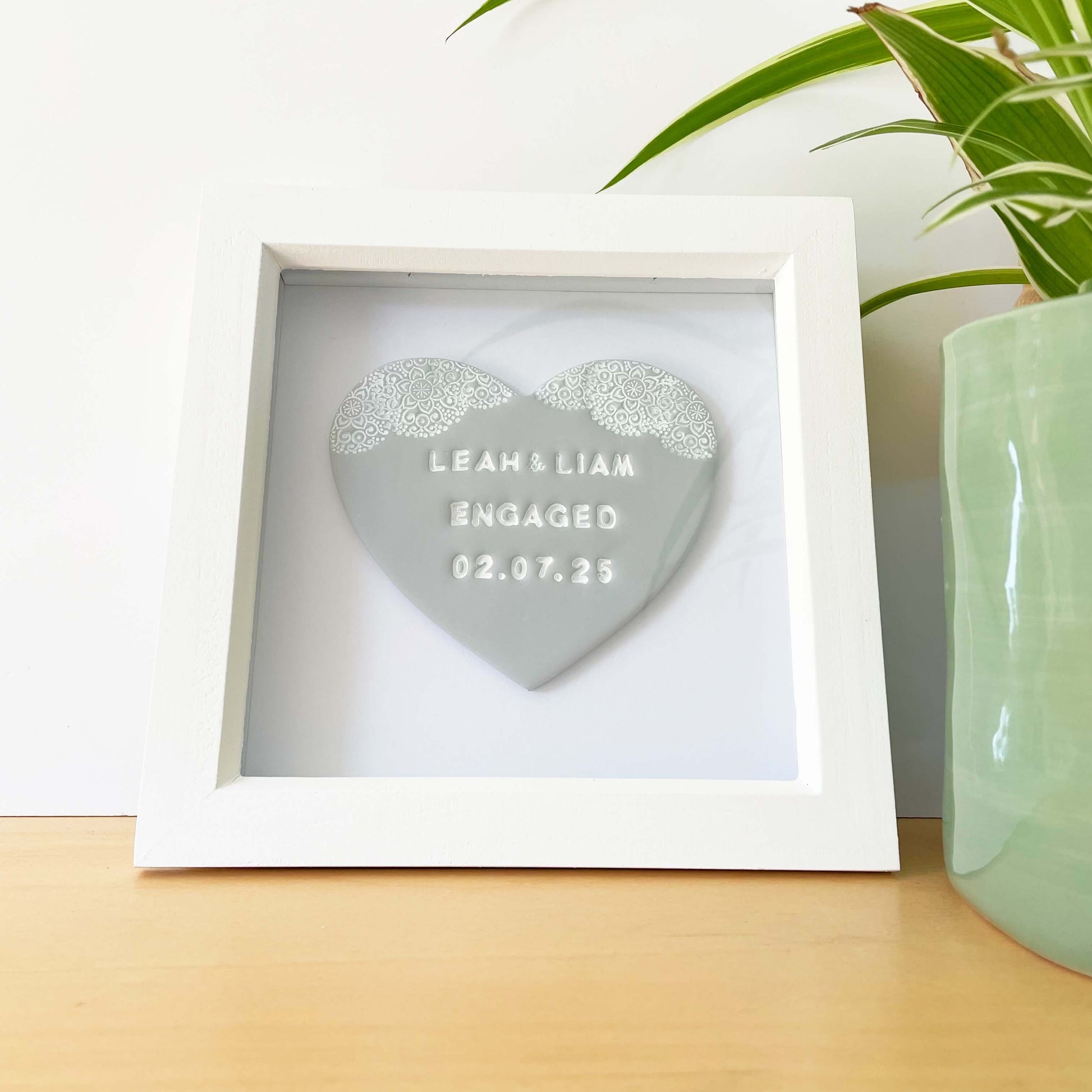 Personalised framed engagement gift, grey clay heart with a white lace edge at the top of the heart in a white box frame, the heart is personalised with LEAH & LIAM ENGAGED 02.07.25