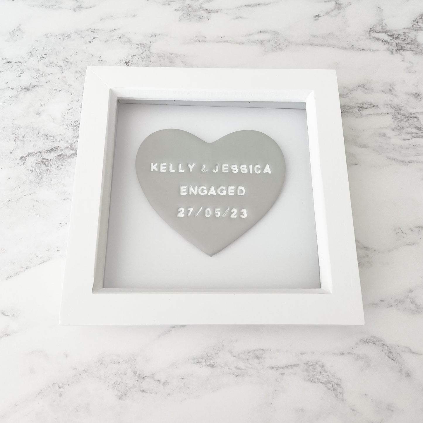 Personalised framed engagement gift, grey clay heart in a white box frame, the heart is personalised with KELLY & JESSICA ENGAGED 27/05/23