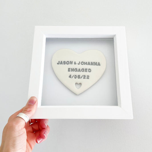 Personalised framed engagement gift, pearlised white clay heart with a heart cut out of the bottom in a white box frame, the heart is personalised in grey with JASON & JOHANNA ENGAGED 4/06/22
