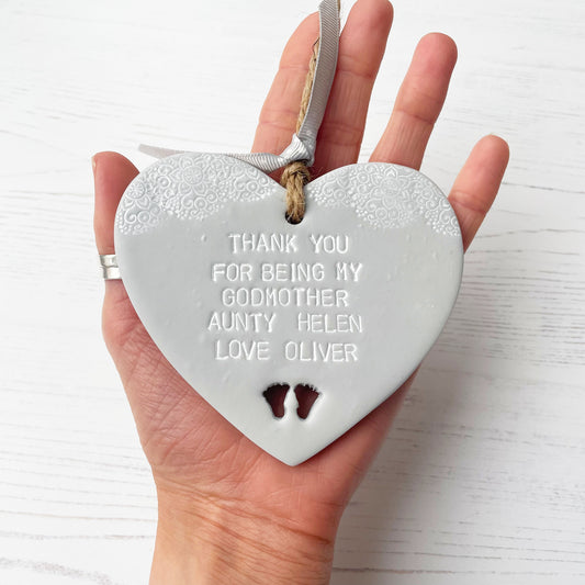 Personalised Godmother gift, grey clay hanging heart with baby feet cut out of the bottom and white lace edge at the top of the heart, the heart is personalised with THANK YOU FOR BEING MY GODMOTHER HELEN LOVE OLIVER 