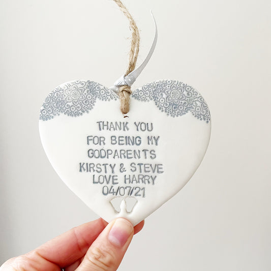 Personalised Godparent gift, pearlised white clay hanging heart with baby feet cut out of the bottom and grey lace edge at the top of the heart, the heart is personalised with THANK YOU FOR BEING MY GODPARENTS KIRSTY & STEVE LOVE HARRY 04/07/21