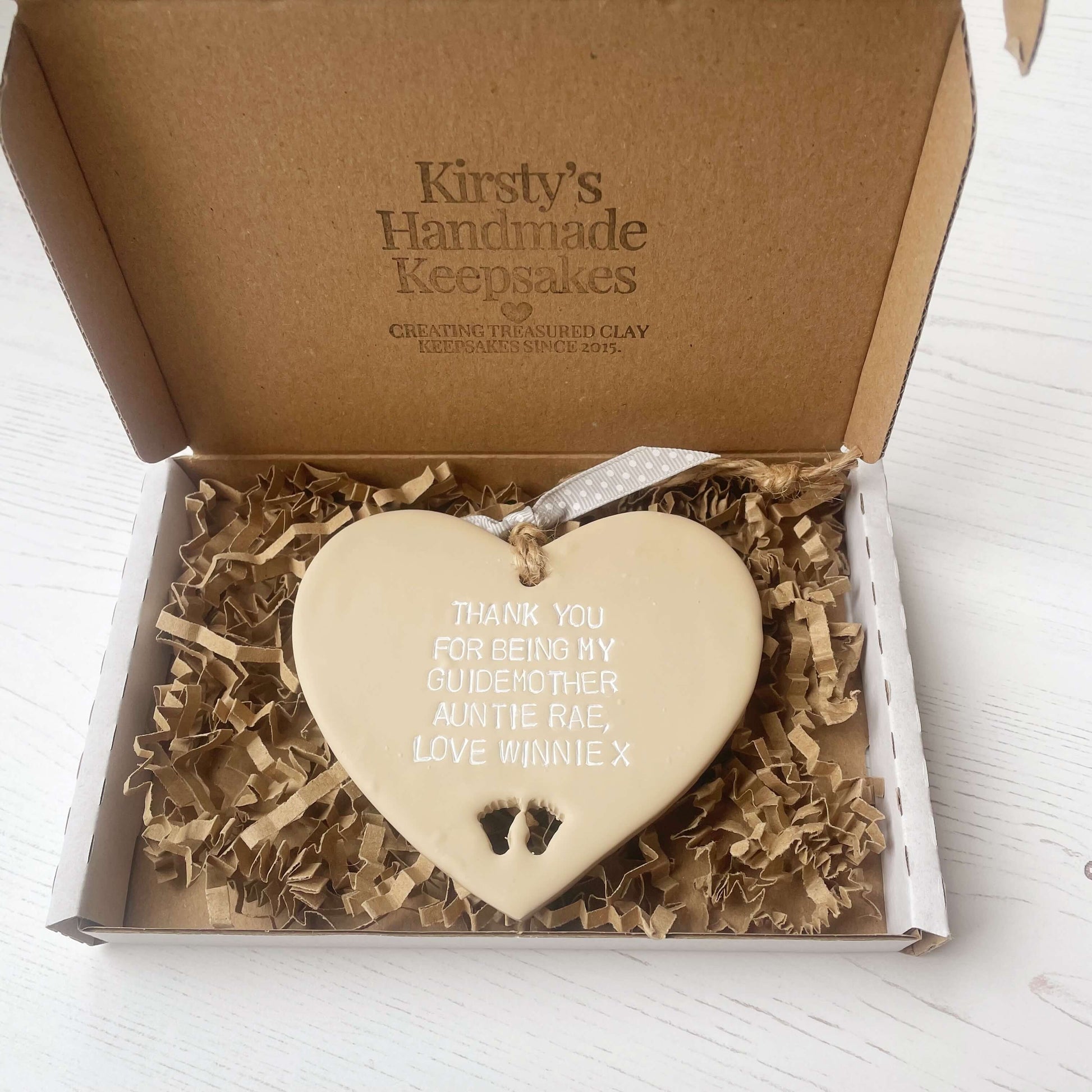 Personalised Guide parent gift, beige clay hanging heart with baby feet cut out of the bottom, the heart is personalised in white with THANK YOU FOR BEING MY GUIDEMOTHER AUNTIE RAE, LOVE WINNIE X