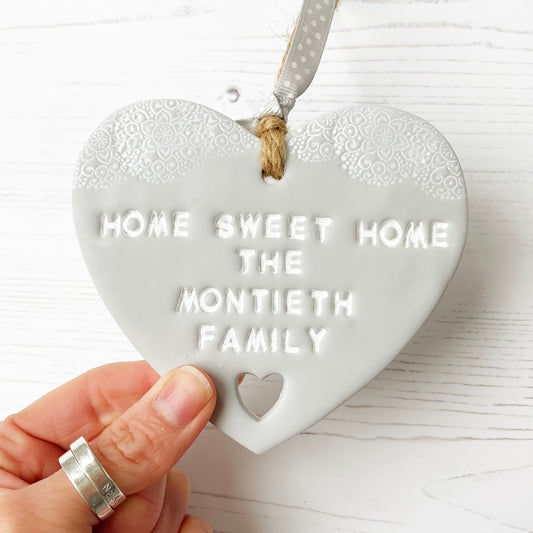 Personalised housewarming new home gift, grey clay heart with a white lace edge at the top of the heart and a heart cut out at the bottom with jute twine for hanging, the heart is personalised with HOME SWEET HOME THE MONTIETH FAMILY