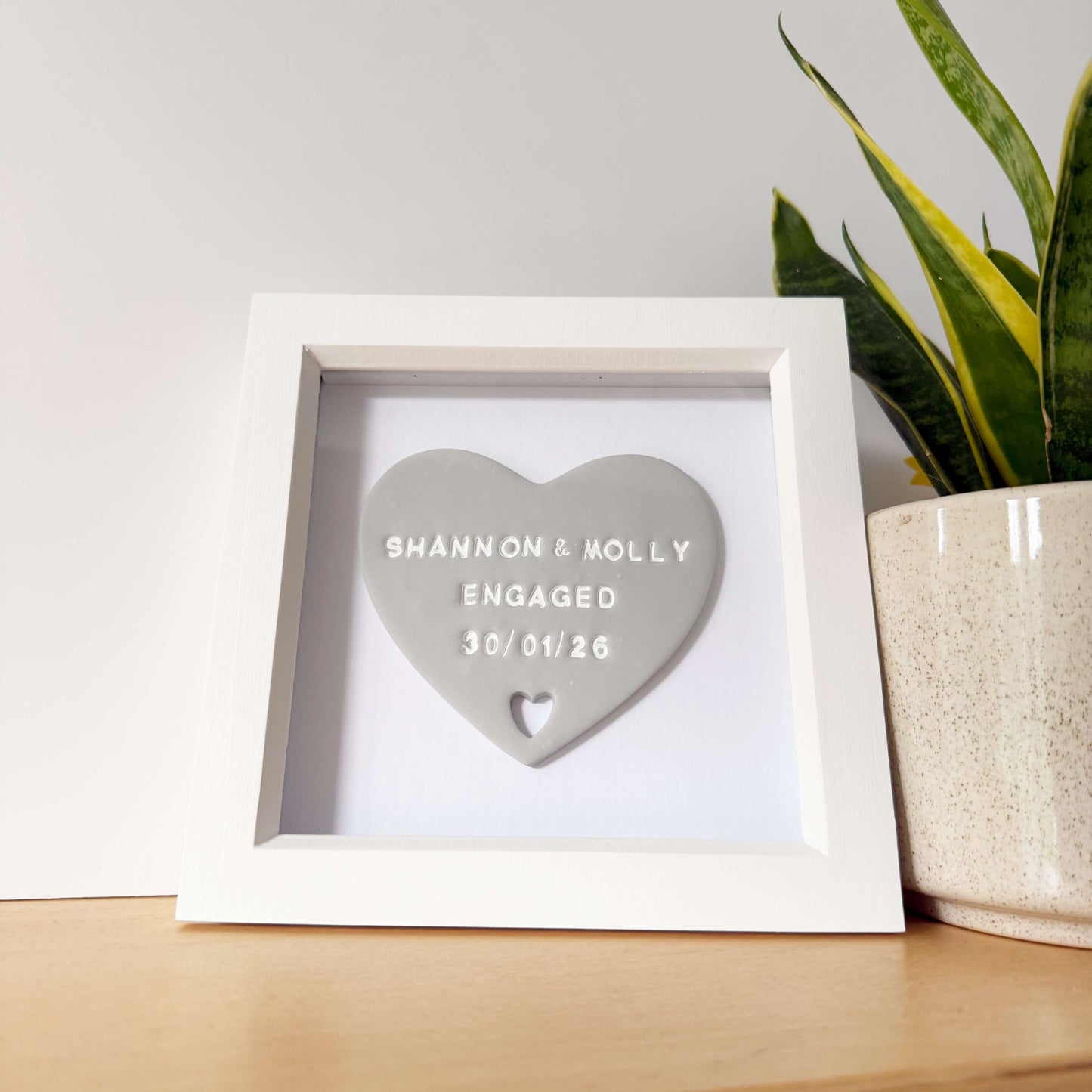 Personalised framed engagement gift, grey clay heart with a heart cut out of the bottom in a white box frame, the heart is personalised in white with SHANNON & MOLLY ENGAGED 30/01/26
