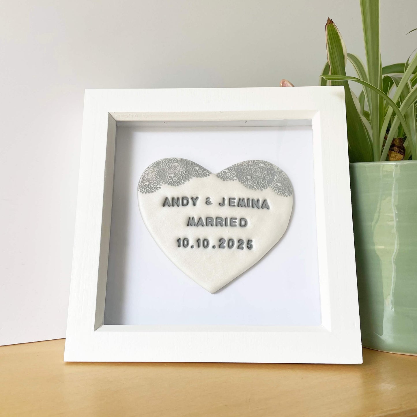Personalised framed wedding gift, pearlised white clay heart with a grey lace edge at the top of the heart in a white box frame, the heart is personalised with ANDY & JEMINA MARRIED 10.10.2025