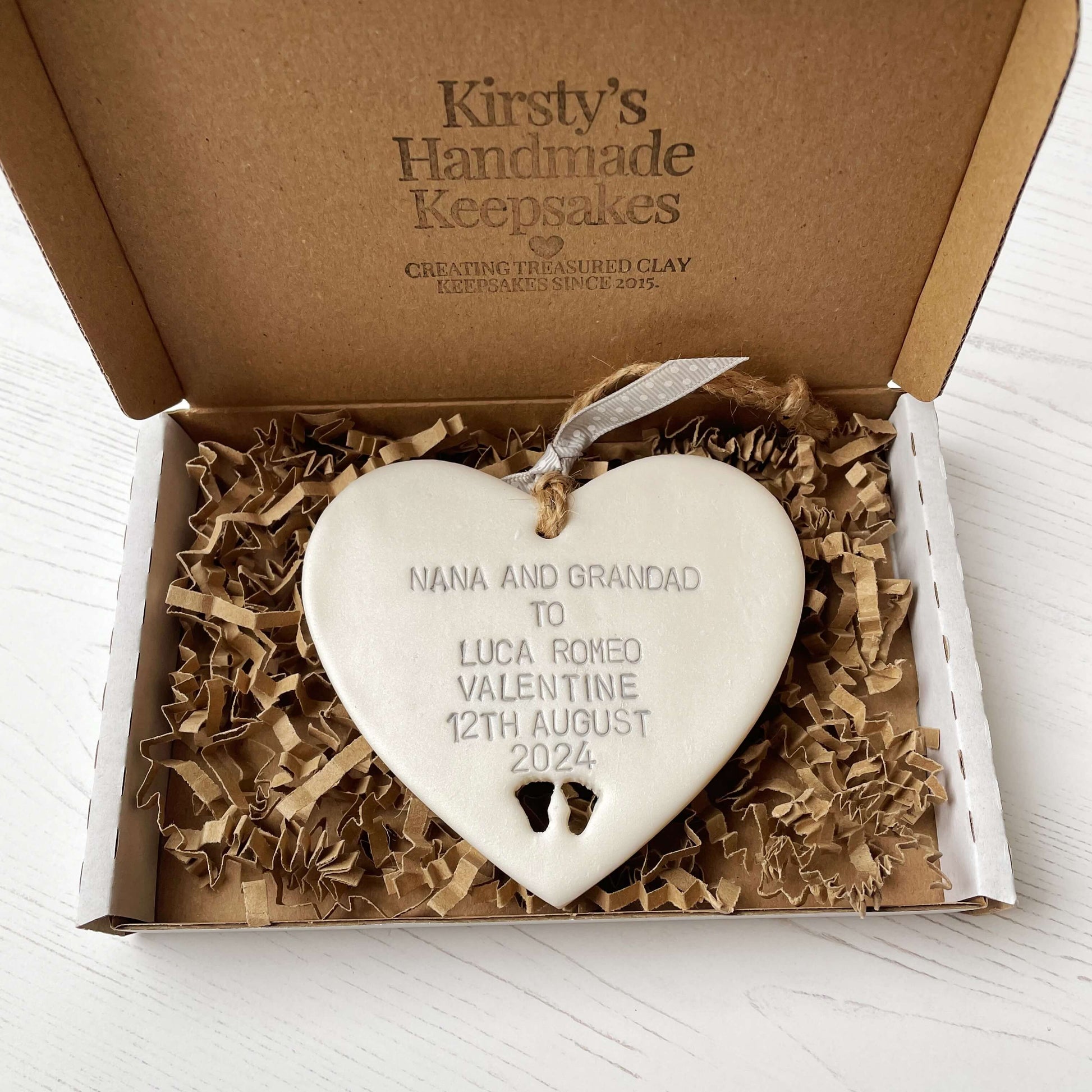 Pearlised white clay hanging heart with baby feet cut out of the bottom and grey personalisation, the heart is personalised with NANA AND GRANDAD TO LUCA ROMEO VALENTINE 12TH AUGUST 2024