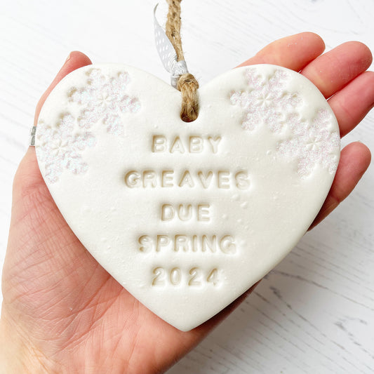 Personalised baby reveal Christmas heart ornament, pearlised white clay with BABY GREAVES DUE SPRING 2024 (text not painted), decorated with 2 iridescent glitter snowflakes on either side of the top of the heart