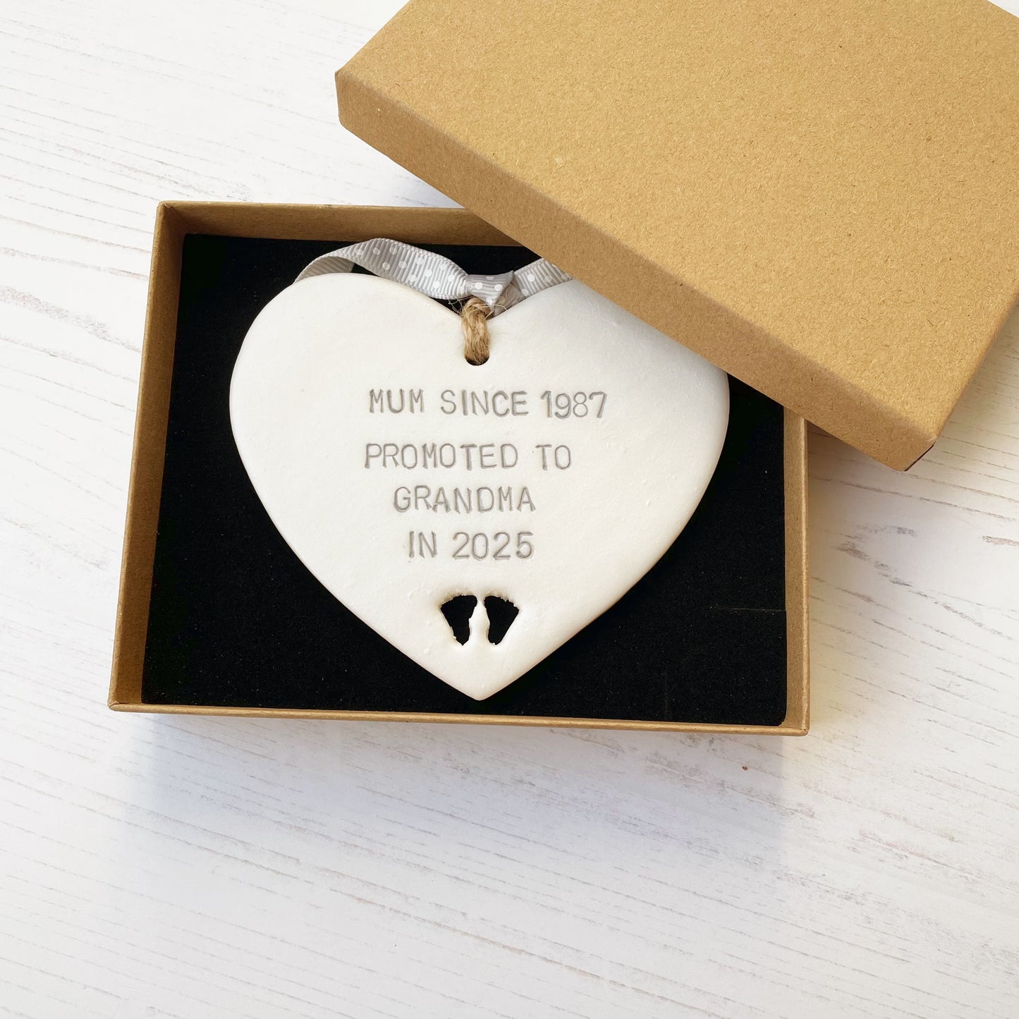 Pearlised white clay hanging heart with baby feet cut out of the bottom and grey personalisation, the heart is personalised with MUM SINCE 1987 PROMOTED TO GRANDMA IN 2025