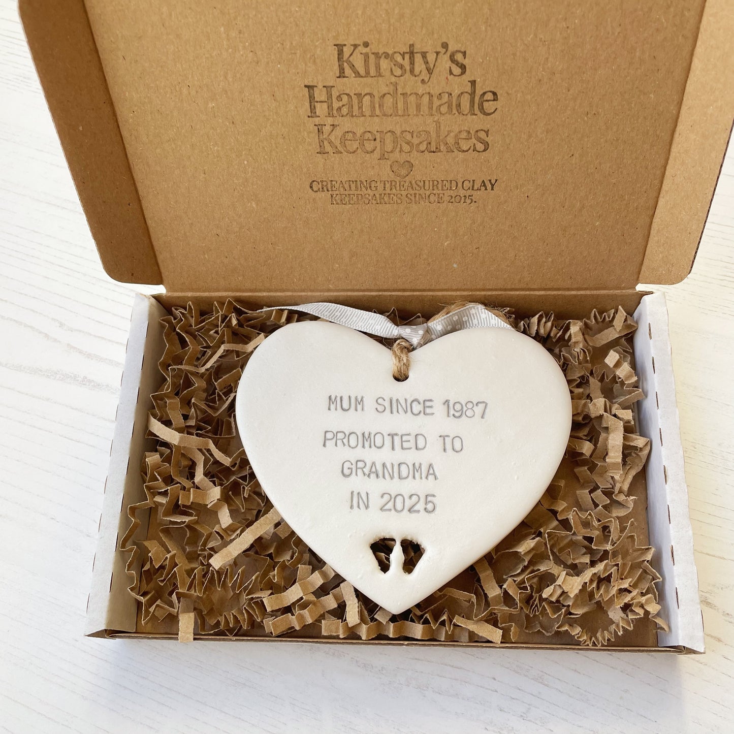 Pearlised white clay hanging heart with baby feet cut out of the bottom and grey personalisation, the heart is personalised with MUM SINCE 1987 PROMOTED TO GRANDMA IN 2025