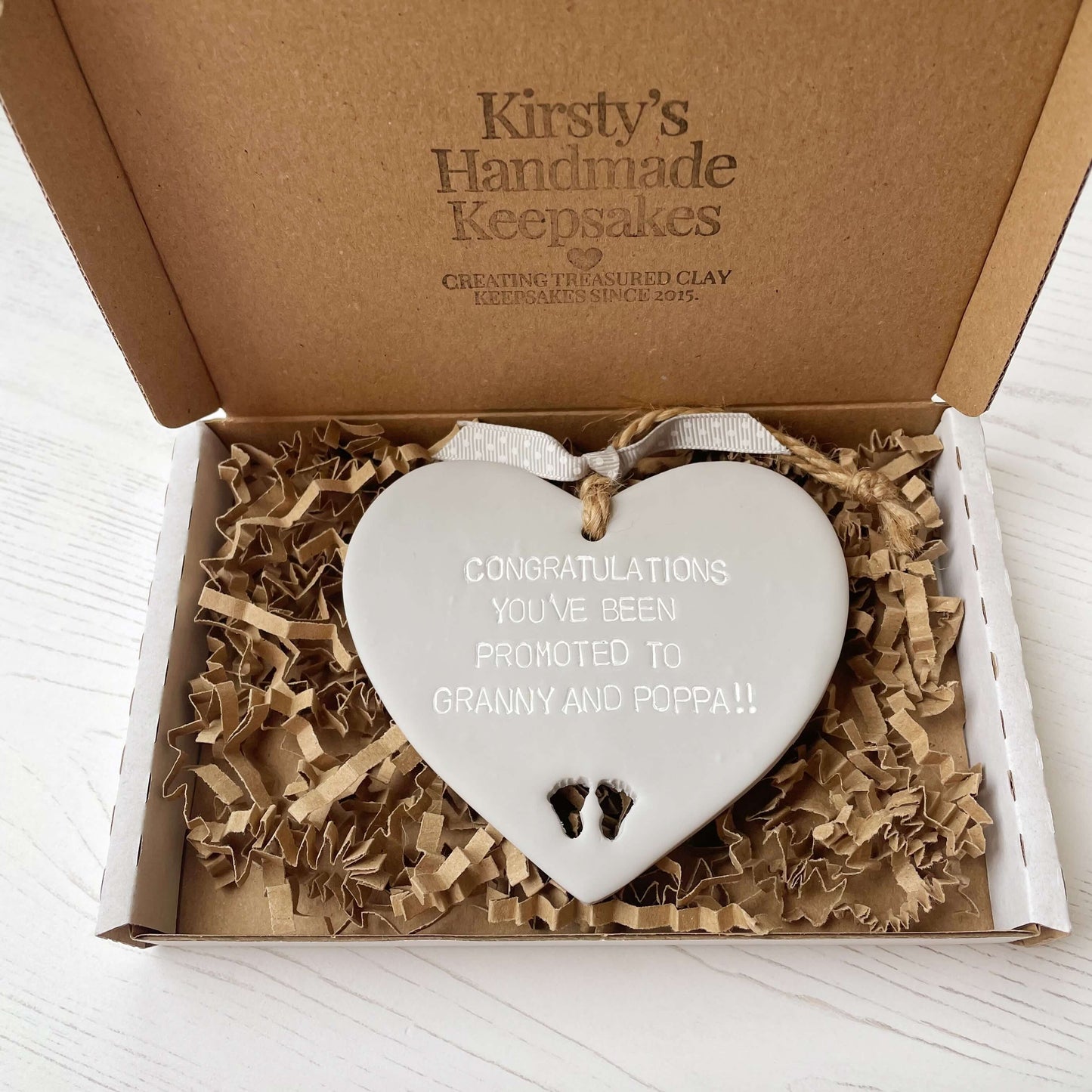 Grey clay hanging heart with baby feet cut out of the bottom and white personalisation, the heart is personalised with CONGRATULATIONS YOU’VE BEEN PROMOTED TO GRANNY AND POPPA!!