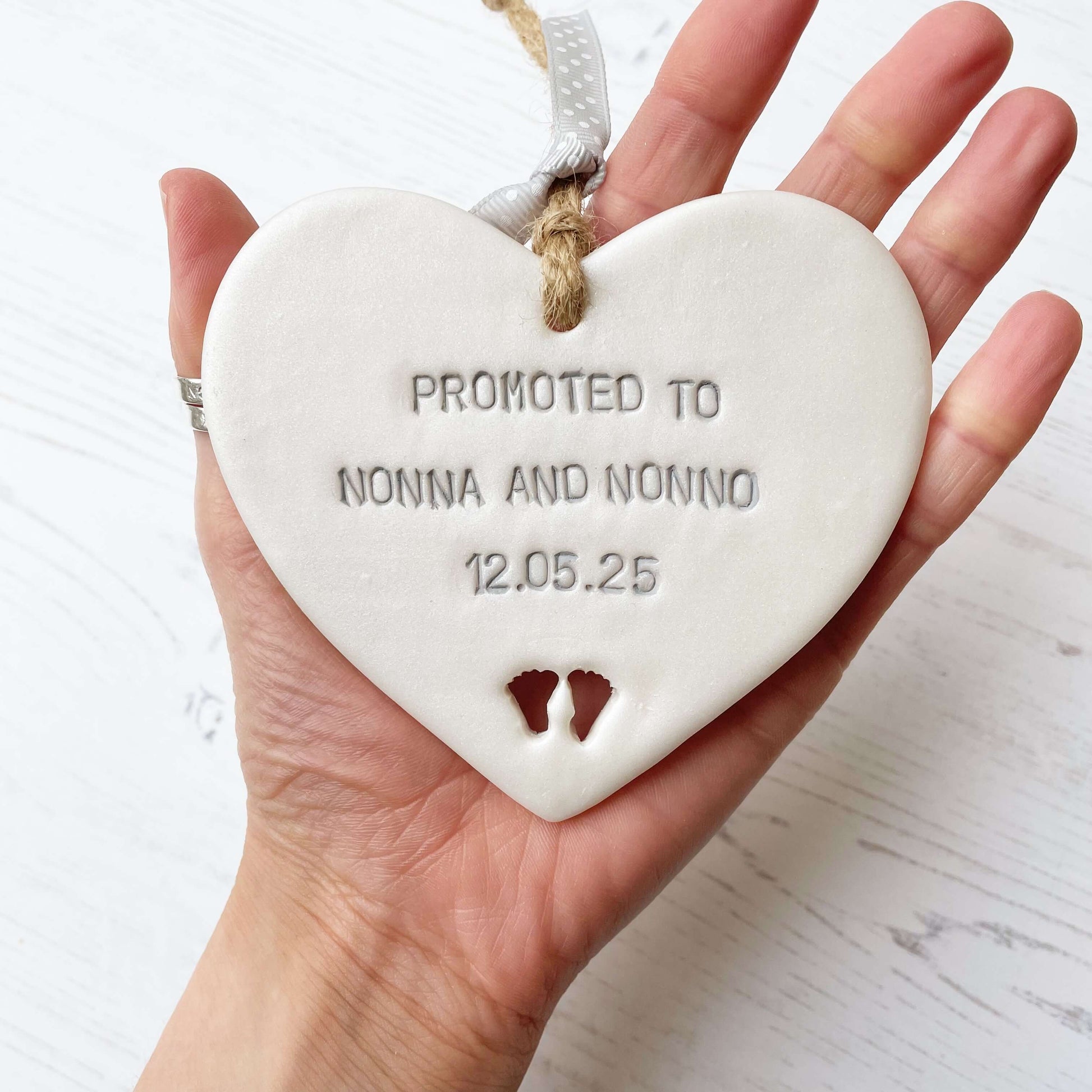 Pearlised white clay hanging heart with baby feet cut out of the bottom and grey personalisation, the heart is personalised with PROMOTED TO NONNA AND NONNO 12.5.25