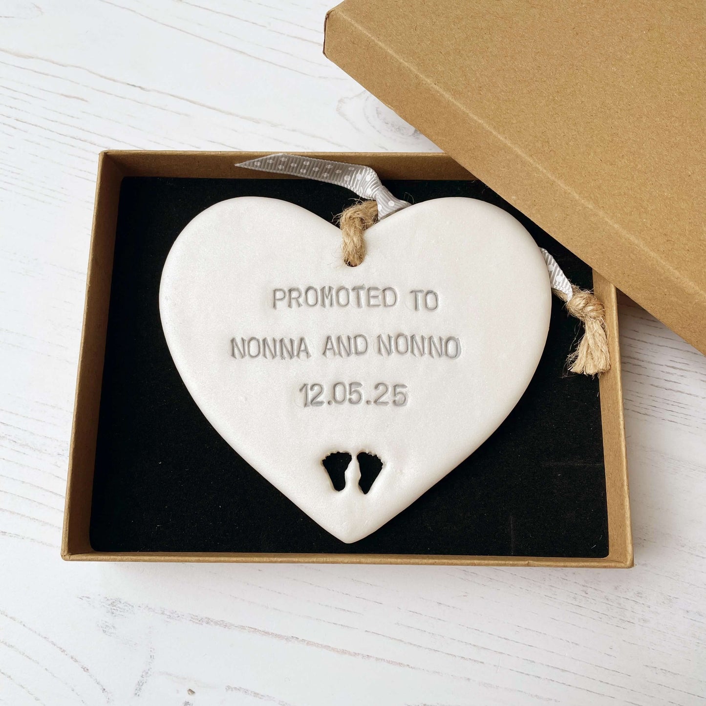 Pearlised white clay hanging heart with baby feet cut out of the bottom and grey personalisation, the heart is personalised with PROMOTED TO NONNA AND NONNO 12.5.25