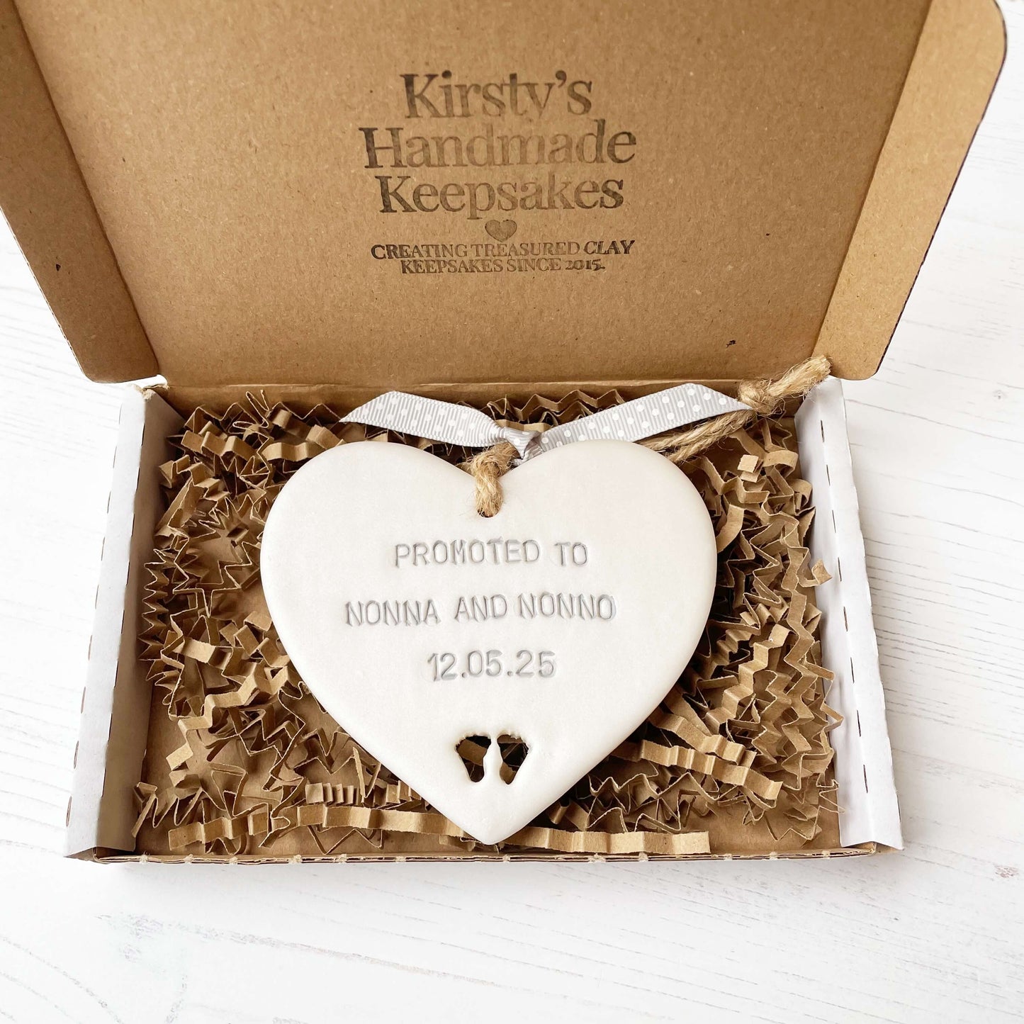 Pearlised white clay hanging heart with baby feet cut out of the bottom and grey personalisation, the heart is personalised with PROMOTED TO NONNA AND NONNO 12.5.25
