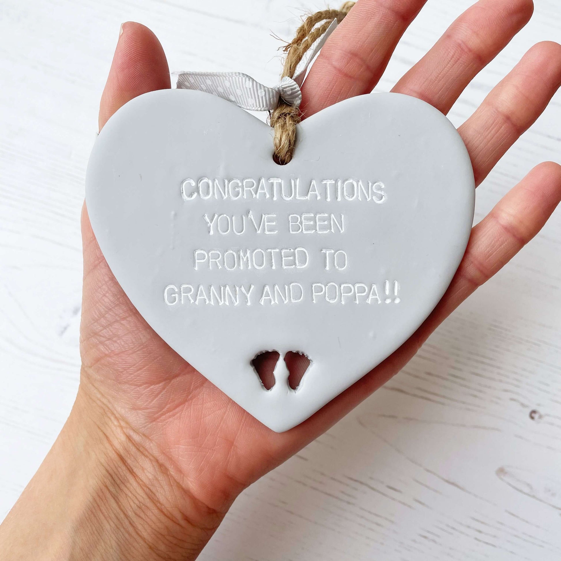 Grey clay hanging heart with baby feet cut out of the bottom and white personalisation, the heart is personalised with CONGRATULATIONS YOU’VE BEEN PROMOTED TO GRANNY AND POPPA!!
