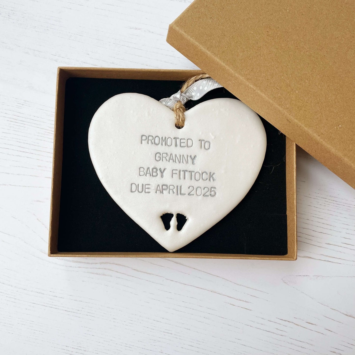 Pearlised white clay hanging heart with baby feet cut out of the bottom and grey personalisation, the heart is personalised with PROMOTED TO GRANNY BABY FITTOCK DUE APRIL 2025