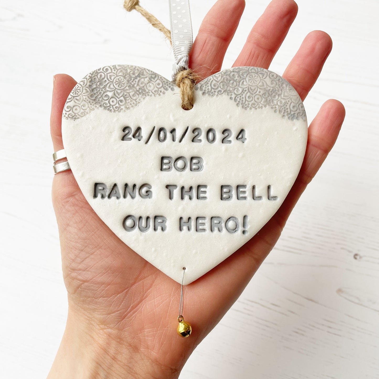 Personalised cancer survivor gift, pearlised white clay hanging heart with a grey lace edge at the top of the heart and a gold bell hanging below, the heart is personalised with 24/01/2024 BOB RANG THE BELL OUR HERO!