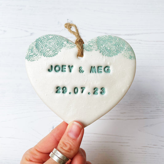 Personalised Valentine’s Day gift, pearlised white clay hanging heart with a sage green lace edge at the top of the heart, the heart is personalised with JOEY & MEG 29.07.23