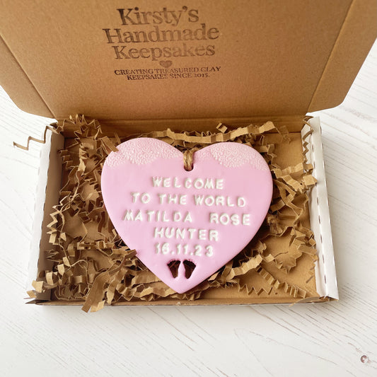 Personalised baby announcement heart sign, in pink clay hanging heart with a white writing and baby feet cut out at the bottom of the heart, the heart is personalised with WELCOME TO THE WORLD BABY MATILDA ROSE HUNTER 16.11.23 In a postal box with brown Kraft shredded zigzag paper