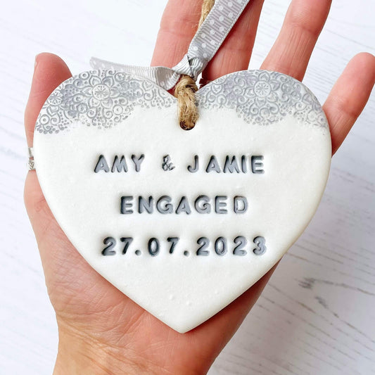 Personalised engagement gift, pearlised white clay heart with a grey lace edge at the top of the heart with jute twine for hanging, the heart is personalised with AMY & JAMIE ENGAGED 27.07.2023