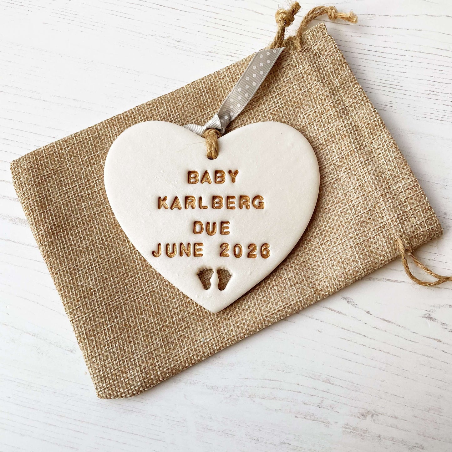 Personalised pregnancy reveal sign keepsake, pearlised white clay hanging heart with baby feet cut out at the bottom, the heart is personalised in gold with BABY KARLBERG DUE JUNE 2026