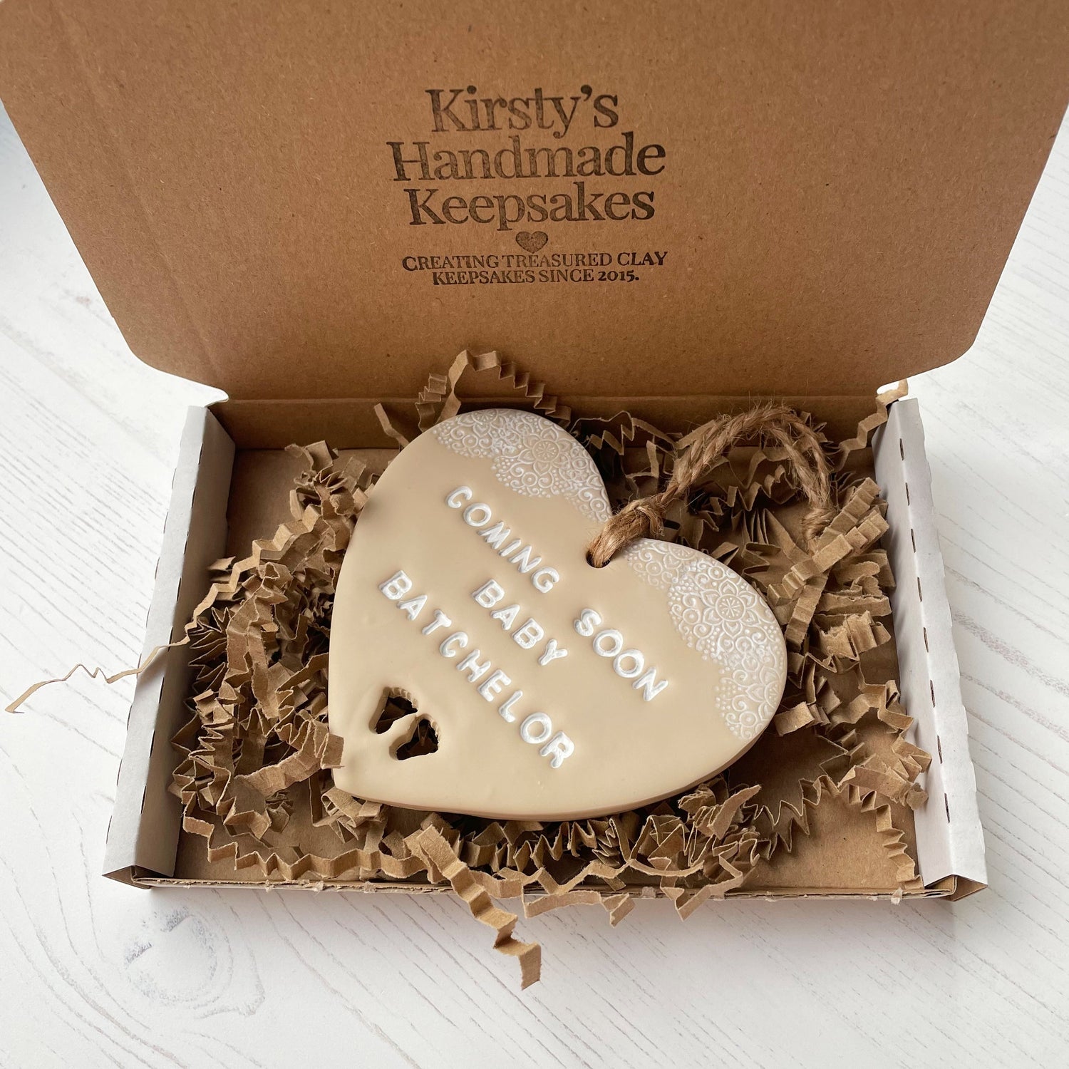 Personalised pregnancy reveal sign keepsake, beige clay hanging heart with a white lace edge at the top of the heart and baby feet cut out at the bottom, the heart is personalised with COMING SOON BABY BATCHELOR. In a white postal box with brown Kraft zigzag shredded paper.