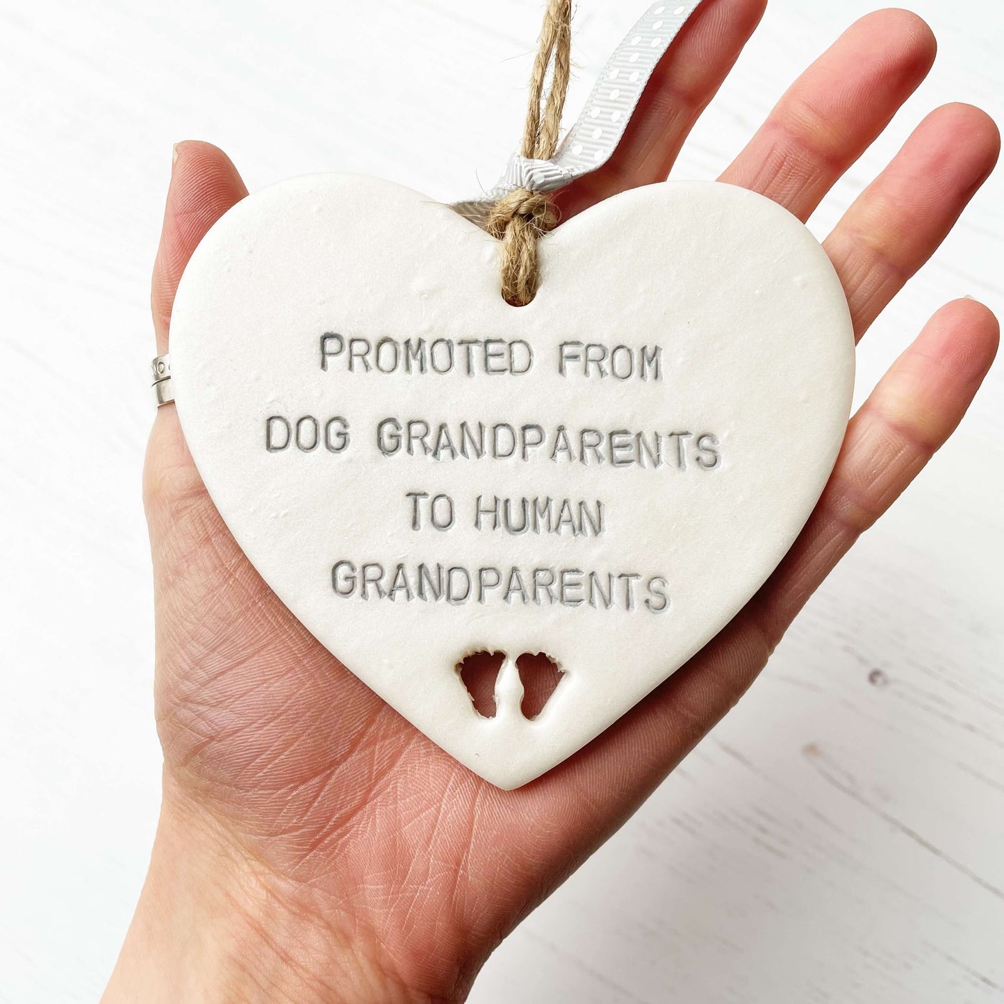 Pearlised white clay hanging heart with baby feet cut out of the bottom and grey personalisation, the heart is personalised with PROMOTED FROM DOG GRANDPARENTS TO HUMAN GRANDPARENTS