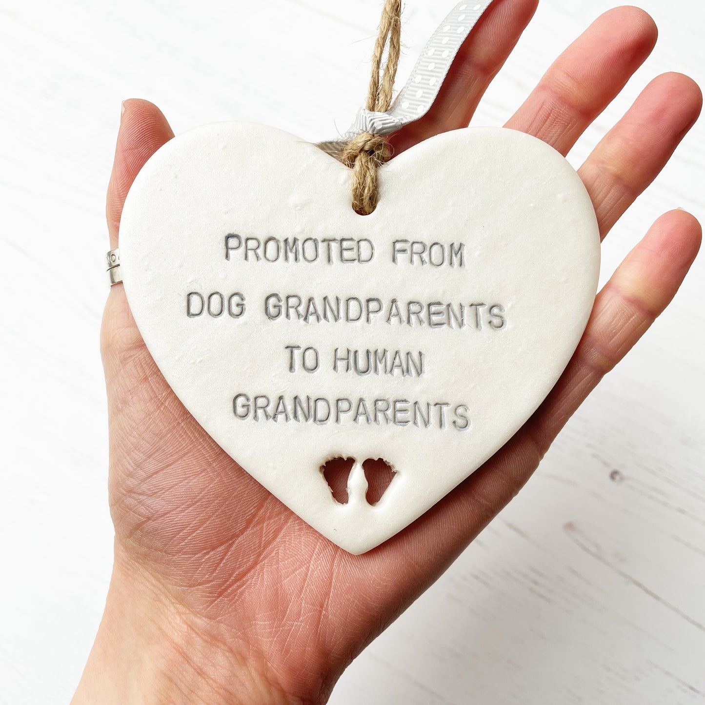 Pearlised white clay hanging heart with baby feet cut out of the bottom and grey personalisation, the heart is personalised with PROMOTED FROM DOG GRANDPARENTS TO HUMAN GRANDPARENTS