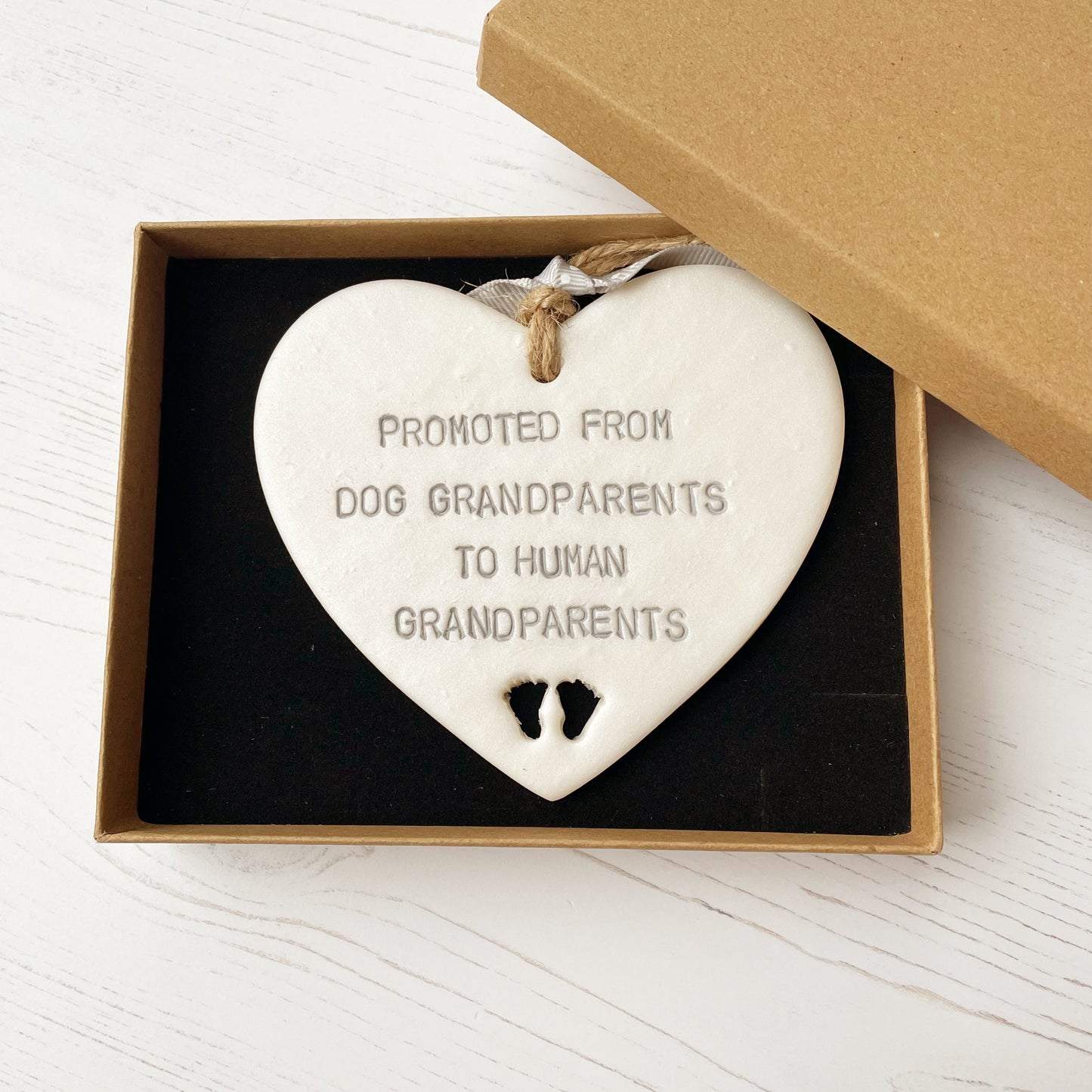 Pearlised white clay hanging heart with baby feet cut out of the bottom and grey personalisation, the heart is personalised with PROMOTED FROM DOG GRANDPARENTS TO HUMAN GRANDPARENTS