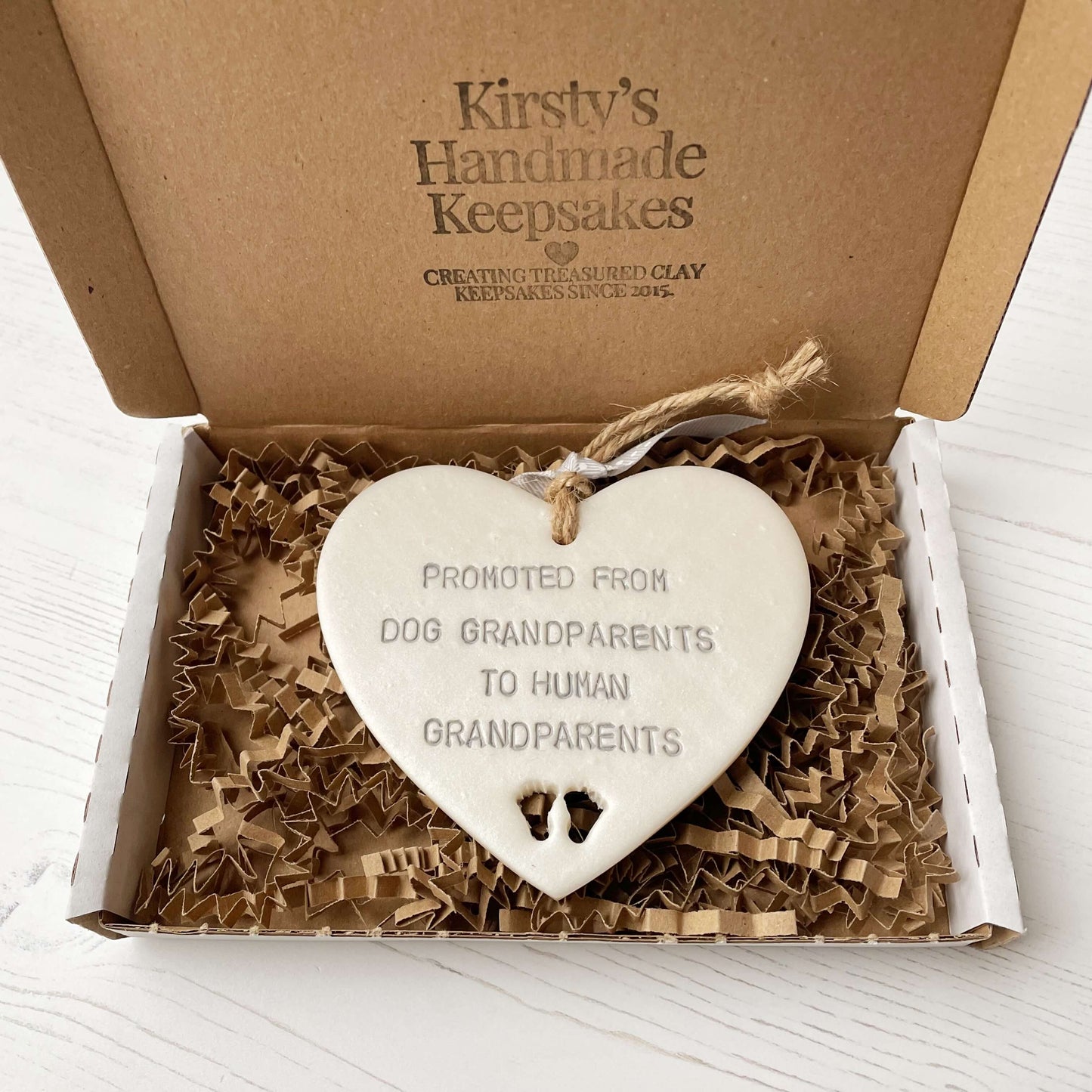 Pearlised white clay hanging heart with baby feet cut out of the bottom and grey personalisation, the heart is personalised with PROMOTED FROM DOG GRANDPARENTS TO HUMAN GRANDPARENTS