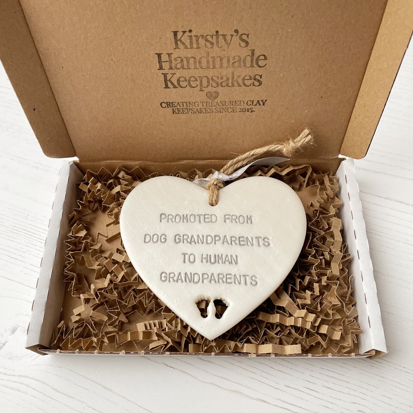 Pearlised white clay hanging heart with baby feet cut out of the bottom and grey personalisation, the heart is personalised with PROMOTED FROM DOG GRANDPARENTS TO HUMAN GRANDPARENTS
