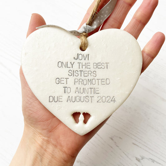 Pearlised white clay hanging heart with baby feet cut out of the bottom and grey personalisation, the heart is personalised with JOVI ONLY THE BEST SISTERS GET PROMOTED TO AUNTIE DUE AUGUST 2024