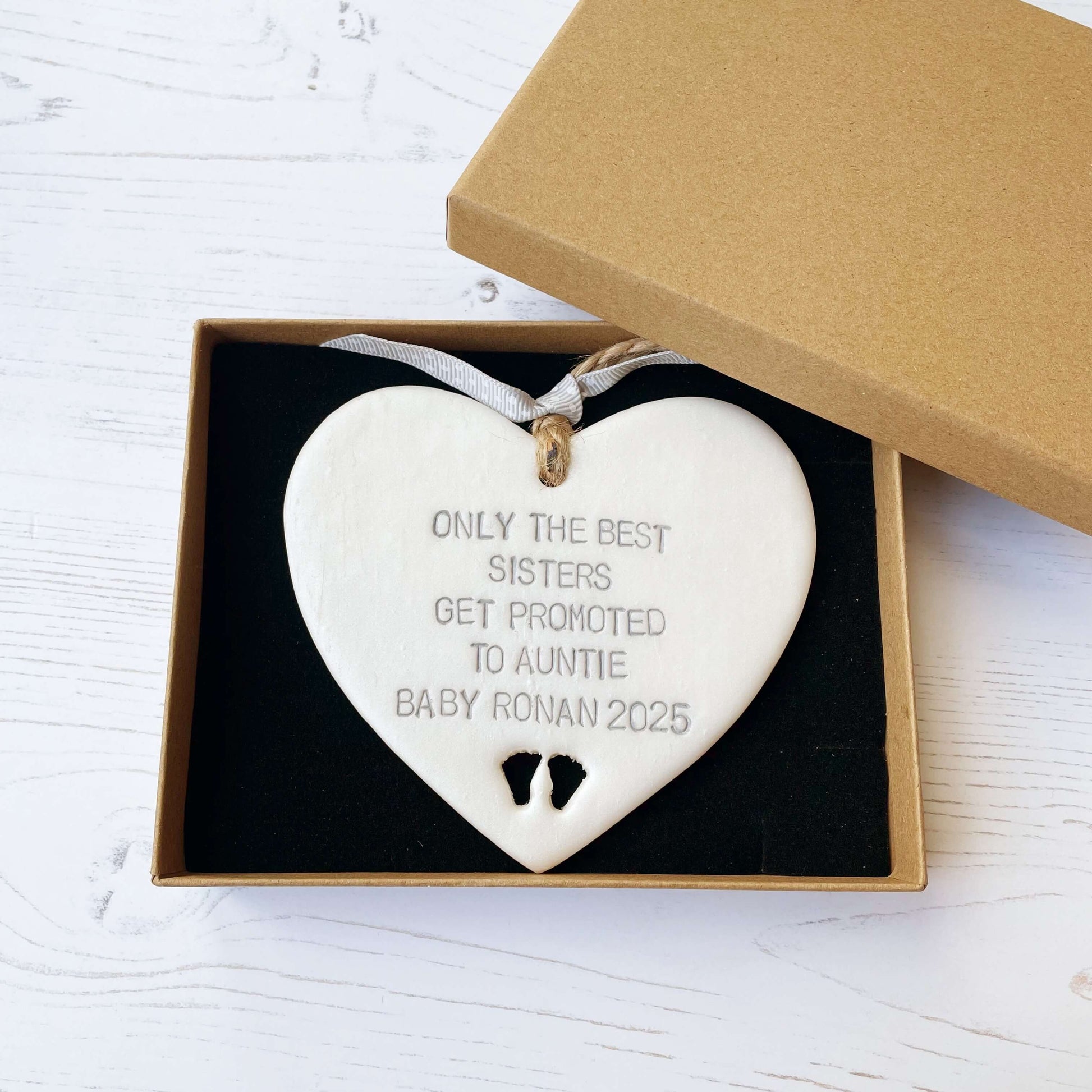 Pearlised white clay hanging heart with baby feet cut out of the bottom and grey personalisation, the heart is personalised with ONLY THE BEST SISTERS GET PROMOTED TO AUNTIE BABY RONAN 2025