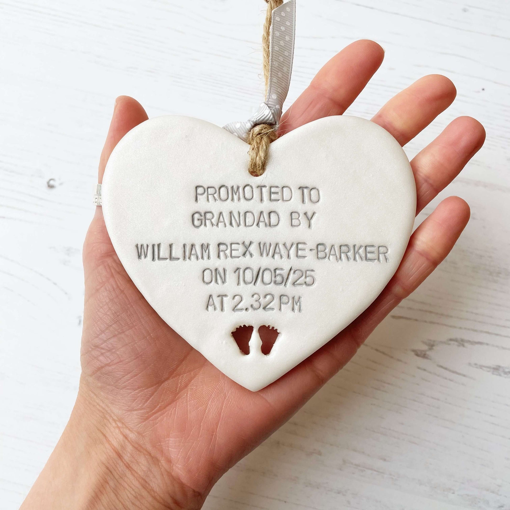 Pearlised white clay hanging heart with baby feet cut out of the bottom and grey personalisation, the heart is personalised with PROMOTED TO GRANDAD BY WILLIAM REX WAYE-BARKER ON 10/05/25 AT 2:32 PM