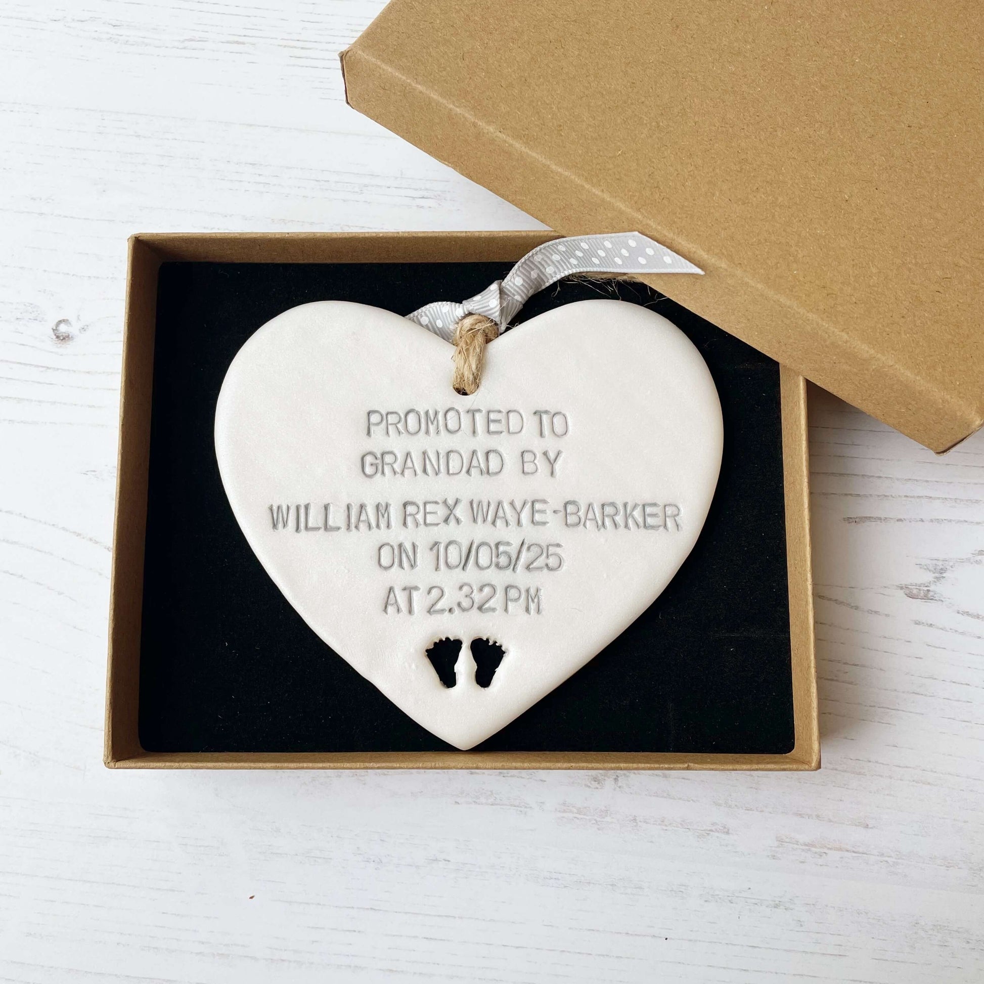 Pearlised white clay hanging heart with baby feet cut out of the bottom and grey personalisation, the heart is personalised with PROMOTED TO GRANDAD BY WILLIAM REX WAYE-BARKER ON 10/05/25 AT 2:32 PM